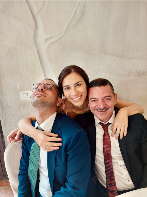 Andrea and Amedeo sitting down with Daniella leaning over, her arms around them.