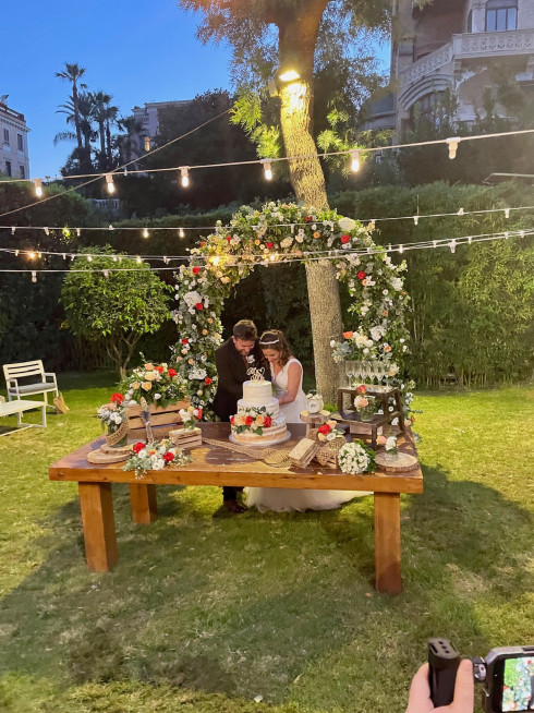 Jay and Filly cut the the cake in the garden, with the wedding arch behind and fairly lights in an evening setting.
